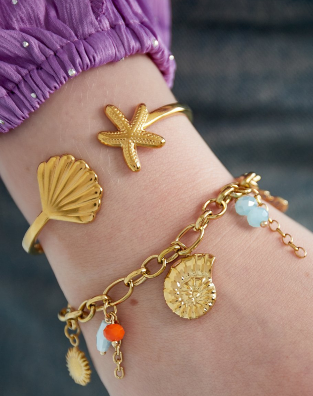 Close-up van een pols met gouden sieraden in een zeethema: een open armband met een zeester en schelp, en een schakelarmband met hangertjes zoals een goudkleurige schelp, zon, en gekleurde kralen. De persoon draagt een paarse blouse met glanzende stipjes.